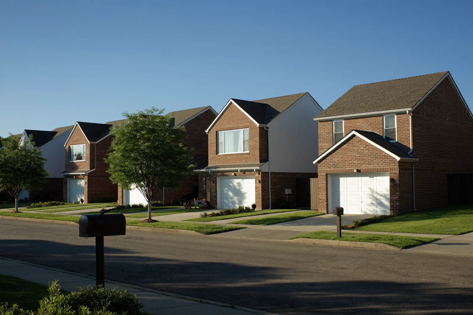 Row of two-story brick homes with white trim and attached garages along a quiet residential street, mailboxes at the kerb in late afternoon sun